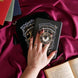 The black Otherworldly clingie by The Quirky Cup Collective is held in front of a matching kindle sticker insert and e-reader sleeve in front of a pink fabric background. The gold kindle grip features an intricate celestial inspired design of moons and stars in a minimal silver, gold, black and white colour palette.