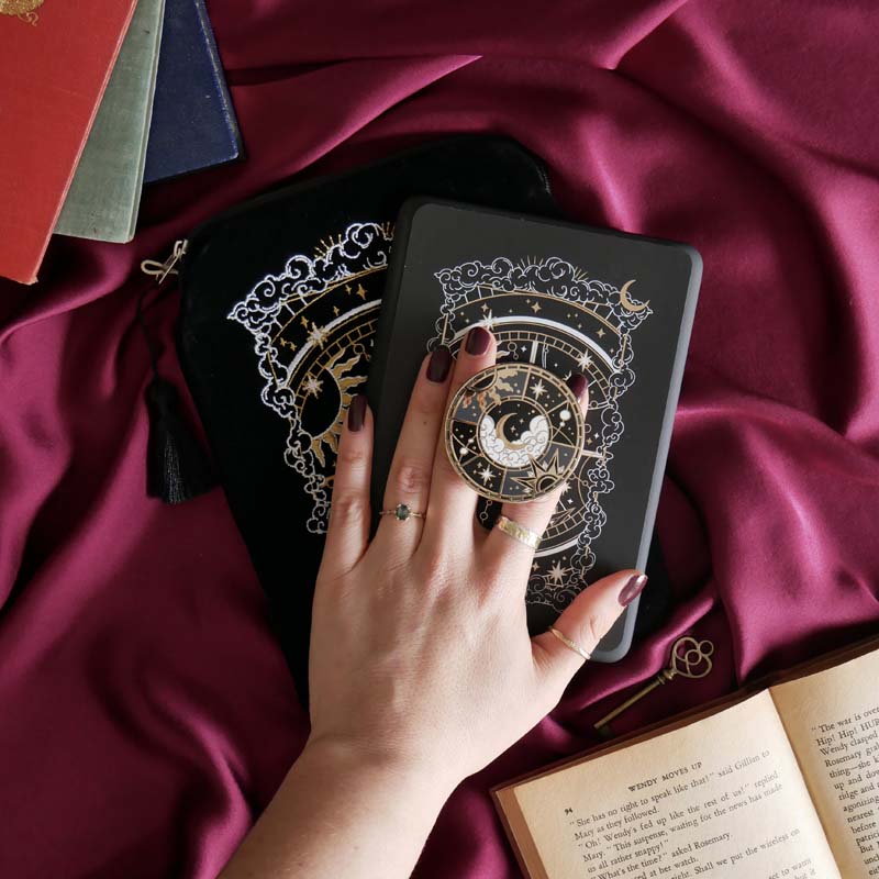 The black Otherworldly clingie by The Quirky Cup Collective is held in front of a matching kindle sticker insert and e-reader sleeve in front of a pink fabric background. The gold kindle grip features an intricate celestial inspired design of moons and stars in a minimal silver, gold, black and white colour palette.