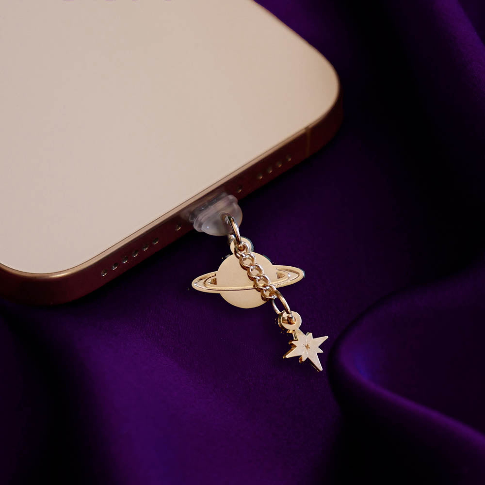 An detail shot of the gold Otherworldly dust plug in a phone on a deep purple fabric background. The book inspired dust plug is feature two charms- a planet wth rings around it and a small star attached via a chain.