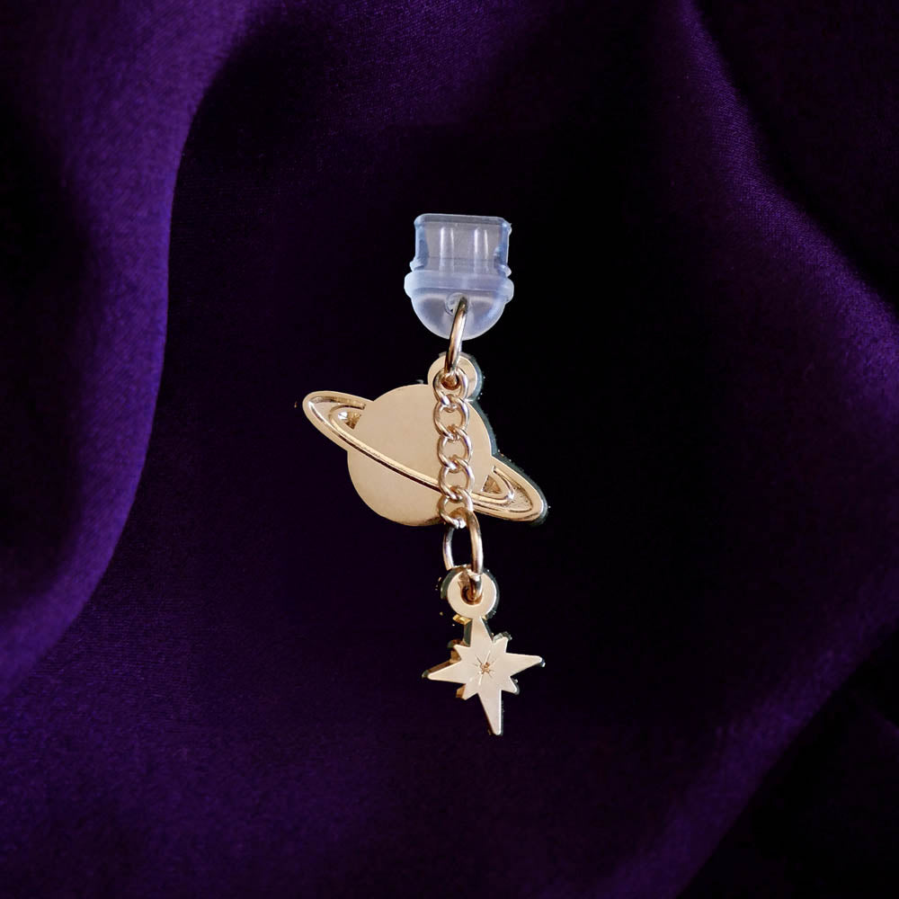 An detail shot of the gold Otherworldly dust plug on a deep purple fabric background. The book inspired dust plug is feature two charms- a planet wth rings around it and a small star attached via a chain.