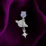 An detail shot of the silver Otherworldly dust plug on a deep purple fabric background. The book inspired dust plug is feature two charms- a planet wth rings around it and a small star attached via a chain.
