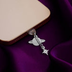 An detail shot of the silver Otherworldly dust plug in a phone on a deep purple fabric background. The book inspired dust plug is feature two charms- a planet wth rings around it and a small star attached via a chain.