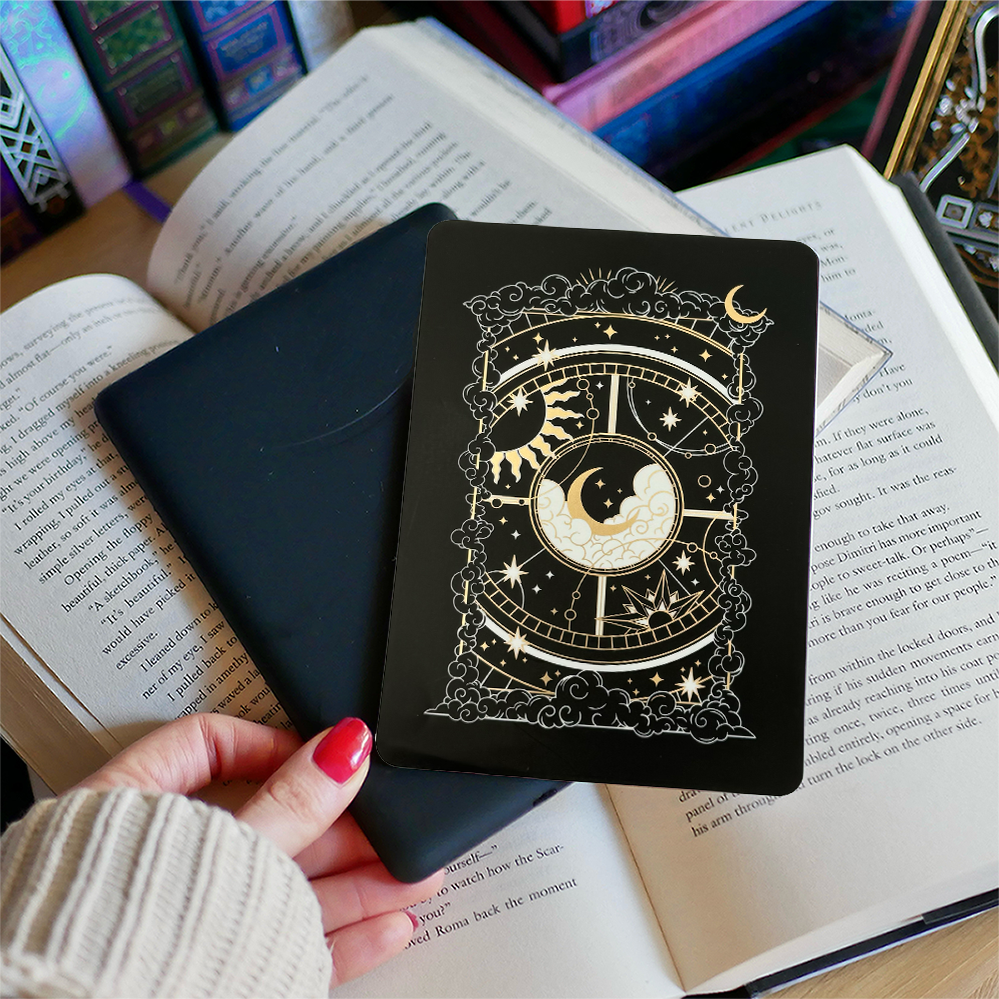 Image shows a hand gently touching a kindle and a kindle sticker insert sitting on top of the device. They sit on a bed of open books. The black, gold and white design shows a moon in the centre, surrounded by a sun, clouds, stars and decorative celestial embellishments. 