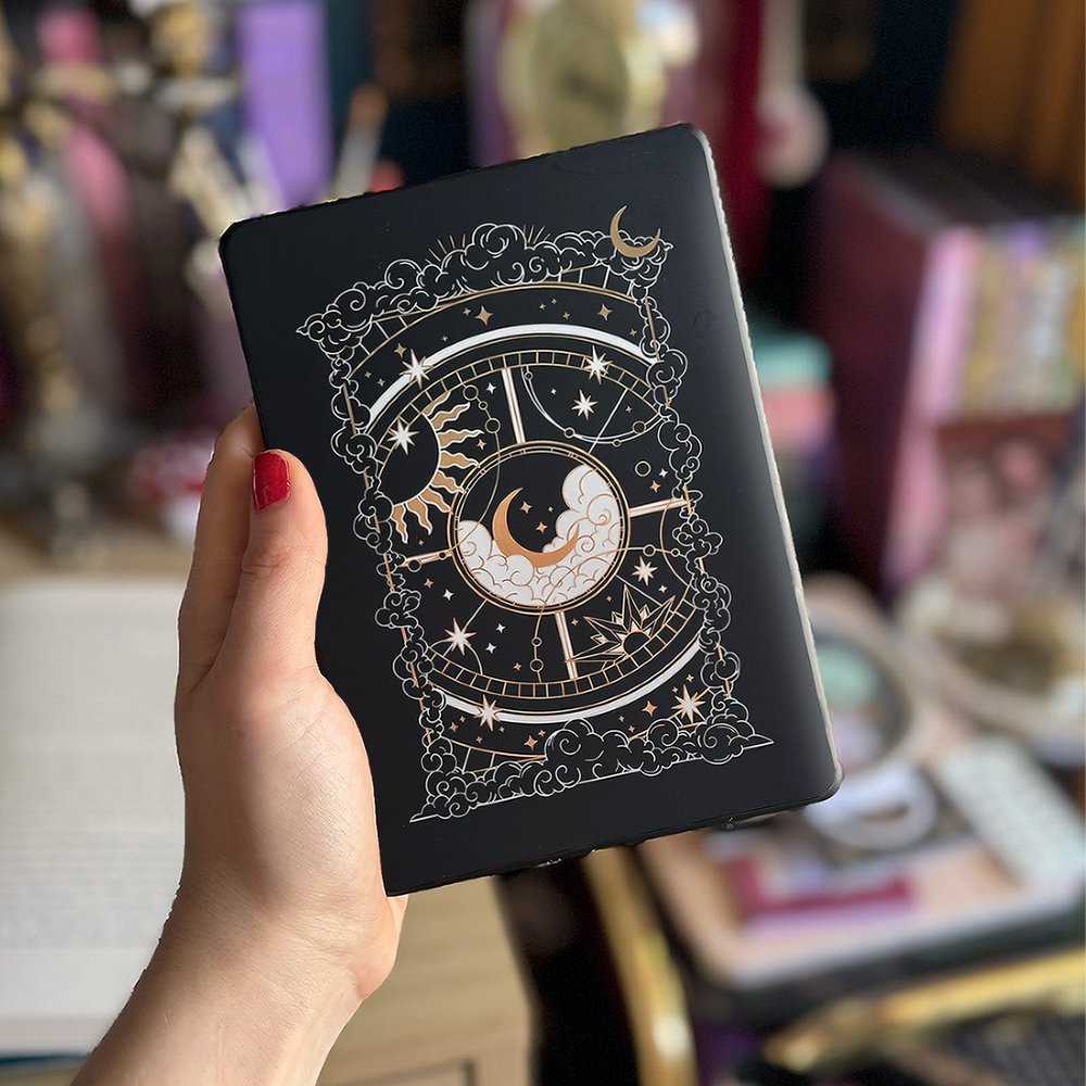 Image shows a hand holding a kindle showing off a kindle sticker insert that is placed on the back of the device. The black, gold and white design shows a moon in the centre, surrounded by a sun, clouds, stars and decorative celestial embellishments. 