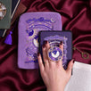 The Otherworldly e-reader bundle set is shown on a pink fabric background and  includes a velvet e-reader case, enamel clingie and kindle sticker insert. The design centers on a celestial moon design surrounded by stars in shades of purple,  the clingie featuring the same design in an circle shape.