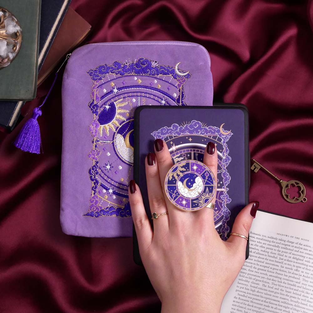 The Otherworldly e-reader bundle set is shown on a pink fabric background and  includes a velvet e-reader case, enamel clingie and kindle sticker insert. The design centers on a celestial moon design surrounded by stars in shades of purple,  the clingie featuring the same design in an circle shape.
