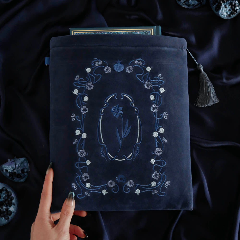 A deep Hades Blue velvet Persephone book sleeve features intricate blue and grey floral embroidery sits on deep blue fabric background. The art nouveau inspired floral design features wheat, skull and pomegranate motifs and the sleeve has a matching blue tassel.