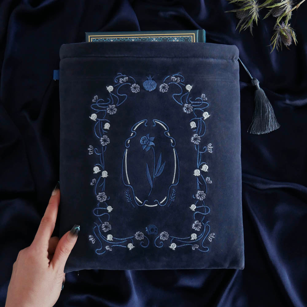 A deep Hades Blue velvet Persephone book sleeve features intricate blue and grey floral embroidery sits on deep blue fabric background. The art nouveau inspired floral design features wheat, skull and pomegranate motifs and the sleeve has a matching blue tassel.