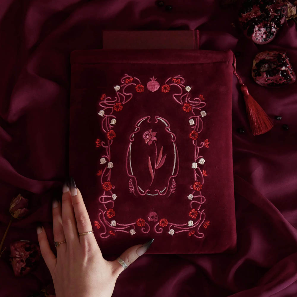 A deep burgundy velvet book sleeve with intricate pink and white floral embroidery, held by a hand over a matching burgundy satin fabric background with dried rose petals and a tassel. Part of the Persephone collection