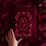 A deep burgundy velvet book sleeve with intricate pink and white floral embroidery, held by a hand over a matching burgundy satin fabric background with dried rose petals and a tassel. Part of the Persephone collection
