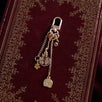 A detail of the Persephone Charmlet (A mix and match accessory for customising your kindle in a romantasy aesthetic) by The Quirky Cup Collective sits on a classical leather bound book in red and gold. The design features a gold clasp and multiple charms hanging from different length detachable chains including a red cut pomegranate, a skull, a flower and a red hart charm.