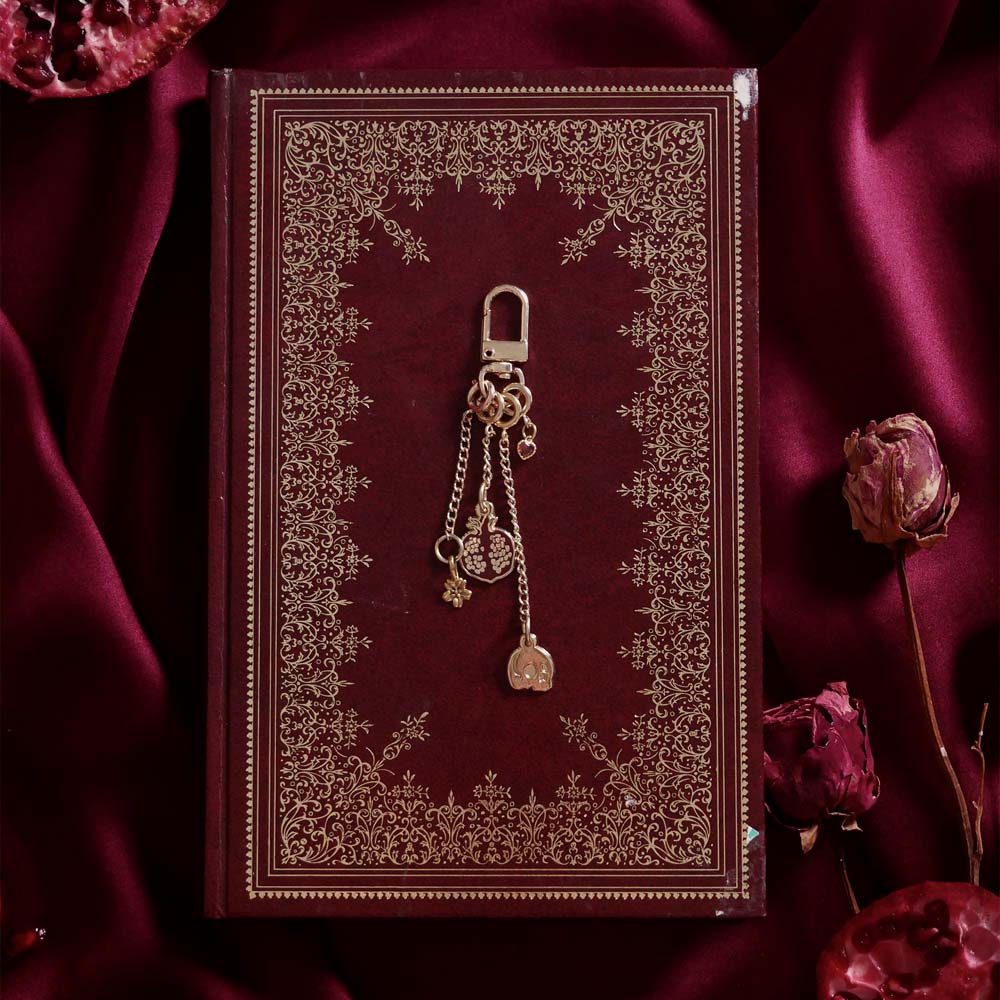 A detail of the Persephone Charmlet (A mix and match accessory for your fantasy inspired kindle aesthetic) by The Quirky Cup Collective sits on a classical leather bound book in red and gold. The design features a gold clasp and multiple charms hanging from different length detachable chains including a red cut pomegranate, a skull, a flower and a red hart charm.
