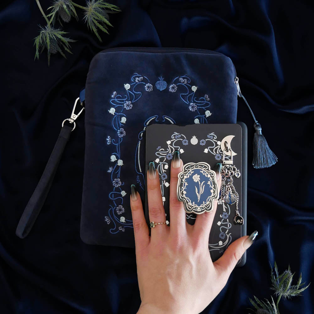 The detail of a hand holding the Hades Blue Persephone eReader bundle is perfect for the classic romance kindle aesthetic. On a blue fabric background sits a Kindle with the Hades Blue version of the Clingie, eReader sticker insert, charmlet (attached with a moon charm anchor) and skull dust plug. A matching velvet eReader sleeve sits under the Kindle.