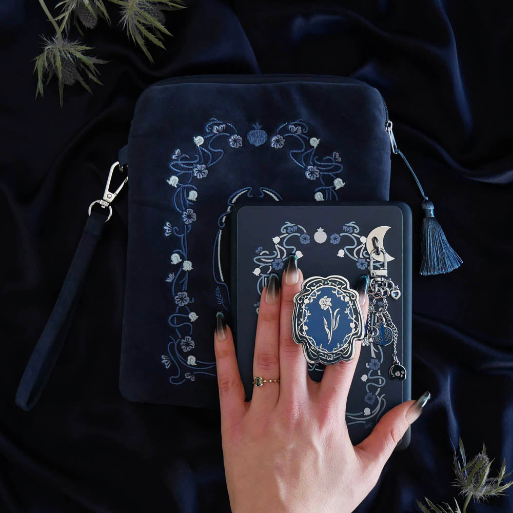 The detail of a hand holding the Hades Blue Persephone eReader bundle is perfect for the classic romance kindle aesthetic. On a blue fabric background sits a Kindle with the Hades Blue version of the Clingie, eReader sticker insert, charmlet (attached with a moon charm anchor) and skull dust plug. A matching velvet eReader sleeve sits under the Kindle.