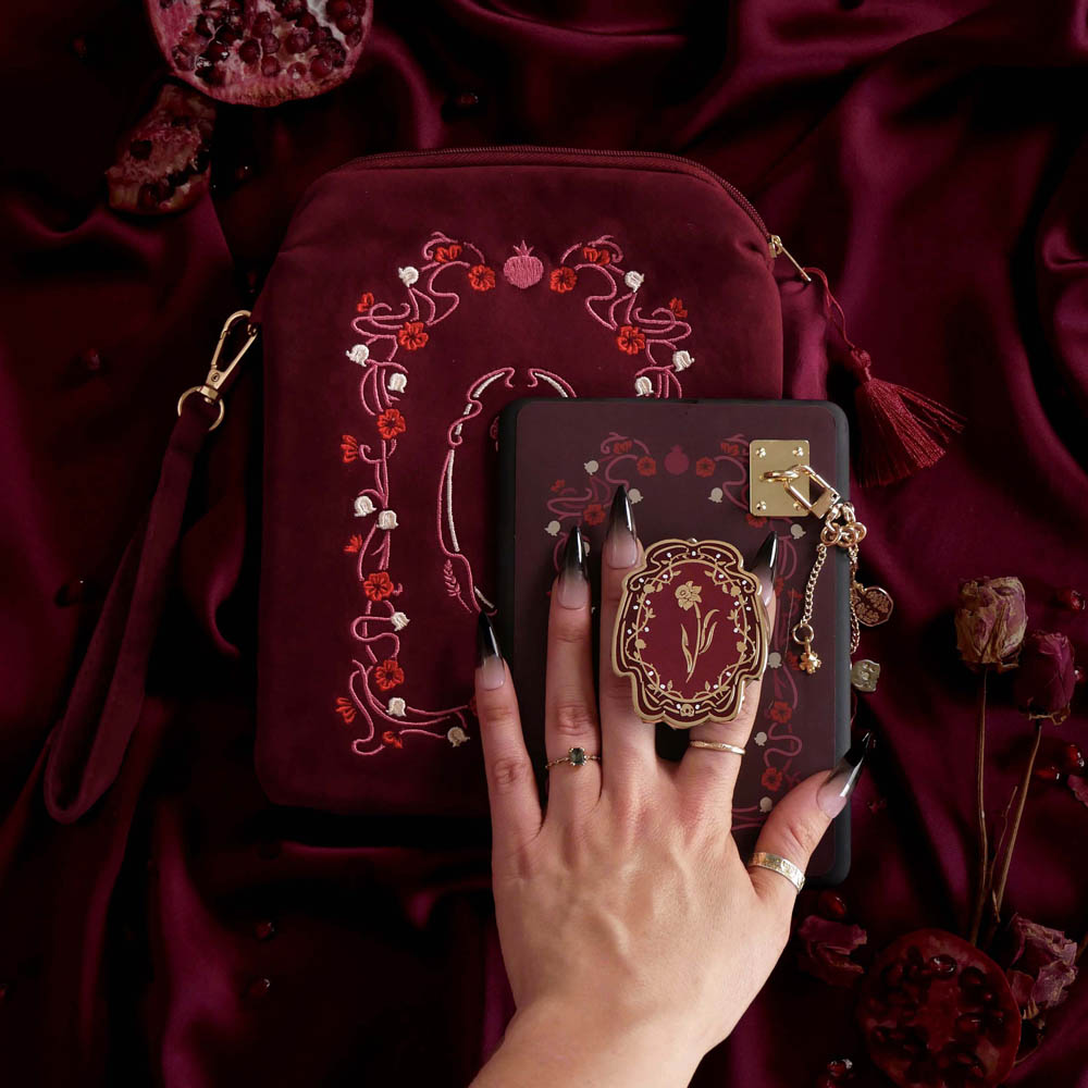 The detail of a hand holding the Wine Persephone eReader bundle is perfect for the classic romance kindle aesthetic. On a deep burgundy fabric background sits a Kindle with the wine red version of the Clingie, eReader sticker insert, charmlet (attached with a rectangle charm anchor) and pomegranate dust plug. A matching velvet eReader sleeve sits under the Kindle.