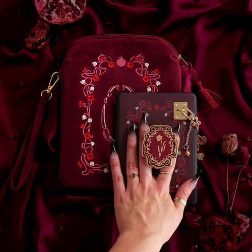 The detail of a hand holding the Wine Persephone eReader bundle is perfect for the classic romance kindle aesthetic. On a deep burgundy fabric background sits a Kindle with the wine red version of the Clingie, eReader sticker insert, charmlet (attached with a rectangle charm anchor) and pomegranate dust plug. A matching velvet eReader sleeve sits under the Kindle.