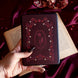 The Persephone e-reader insert sticker is applied to a kindle is held by a hand with long black nails on a blood red fabric background surrounded by roses and pomegranates. The design is in shades of pink, red and cream. The design draws on an art nouveau aesthetic and features a floral frame and design alongside pomegranate and skull motifs.