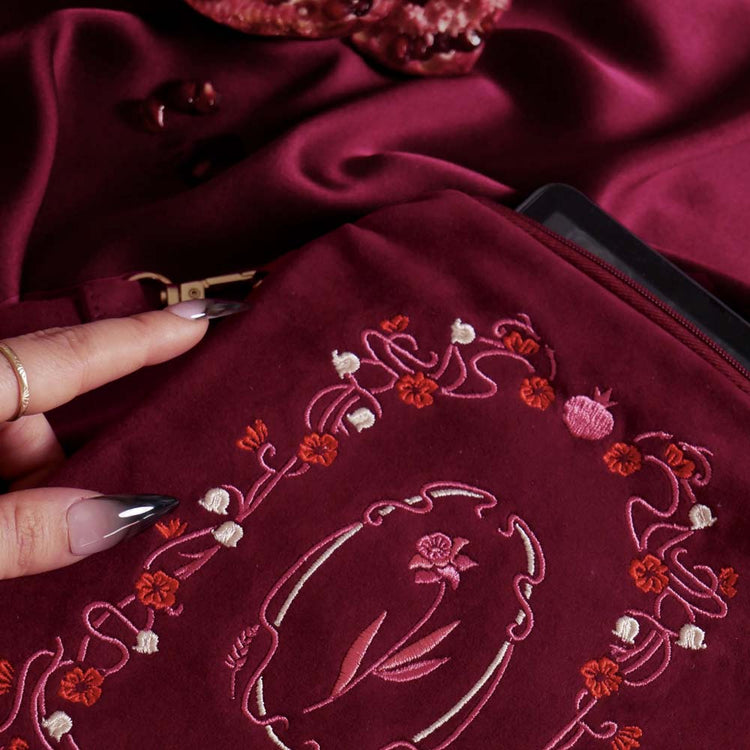 A detail of the Persephone embroidered e-reader case is held by a hand with long black nails on a blood red fabric background surrounded by roses and pomegranates. The kindle case features a wrist strap and is a red velvet colour with pink, red and cream embroidery. The design draws on an art nouveau aesthetic and features a floral frame and design alongside pomegranate and skull motifs.