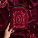 The Persephone embroidered e-reader case is held by a hand with long black nails on a blood red fabric background surrounded by roses and pomegranates. The kindle case features a wrist strap and is a red velvet colour with pink, red and cream embroidery. The design draws on an art nouveau aesthetic and features a floral frame and design alongside pomegranate and skull motifs.