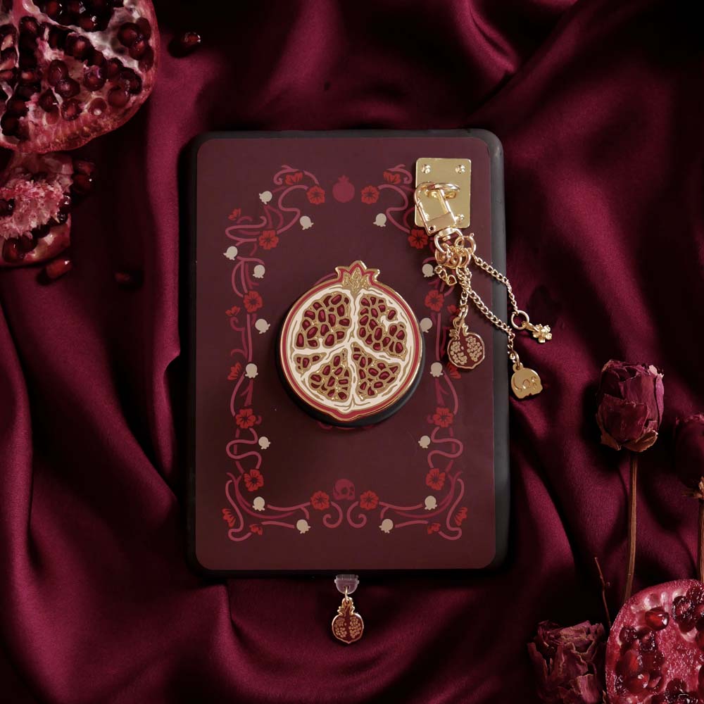 A close up of the Persephone kindle decor bundle by The Quirky Cup Collective featuring a kindle insert sticker, dust plug, charmlet and charm anchor and cling (it's like a bookish pop socket or enamel phone grip!). These romance book inspired kindle products are inspired by the story of Persephone and feature a deep blood red colour alongside motifs of art nouveu flower frames, pomegranates, skulls and flowers.