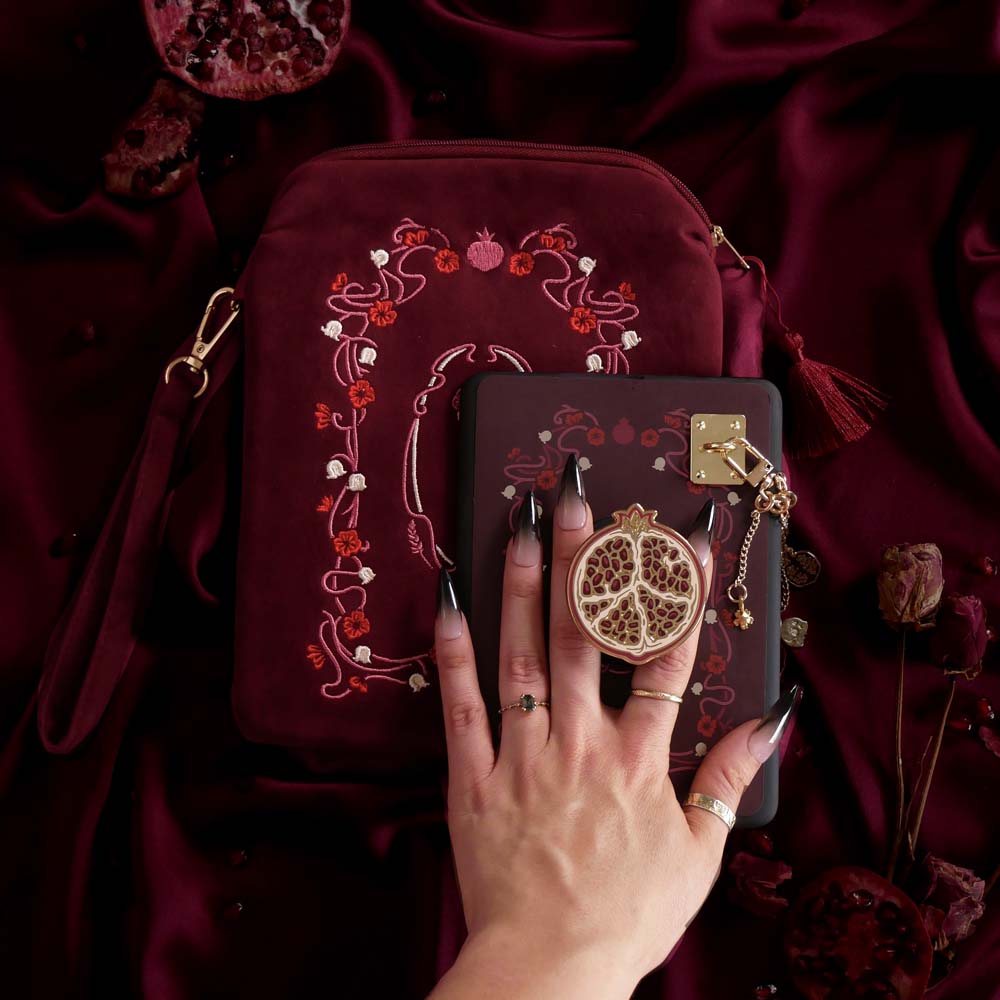 A close up of the Persephone kindle decor bundle by The Quirky Cup Collective featuring a kindle insert sticker, e-reader case, charmlet and charm anchor and cling (it's like a bookish pop socket or enamel phone grip!). These romance book inspired kindle products are inspired by the story of Persephone and feature a deep blood red colour alongside motifs of art nouveu flower frames, pomegranates, skulls and flowers.