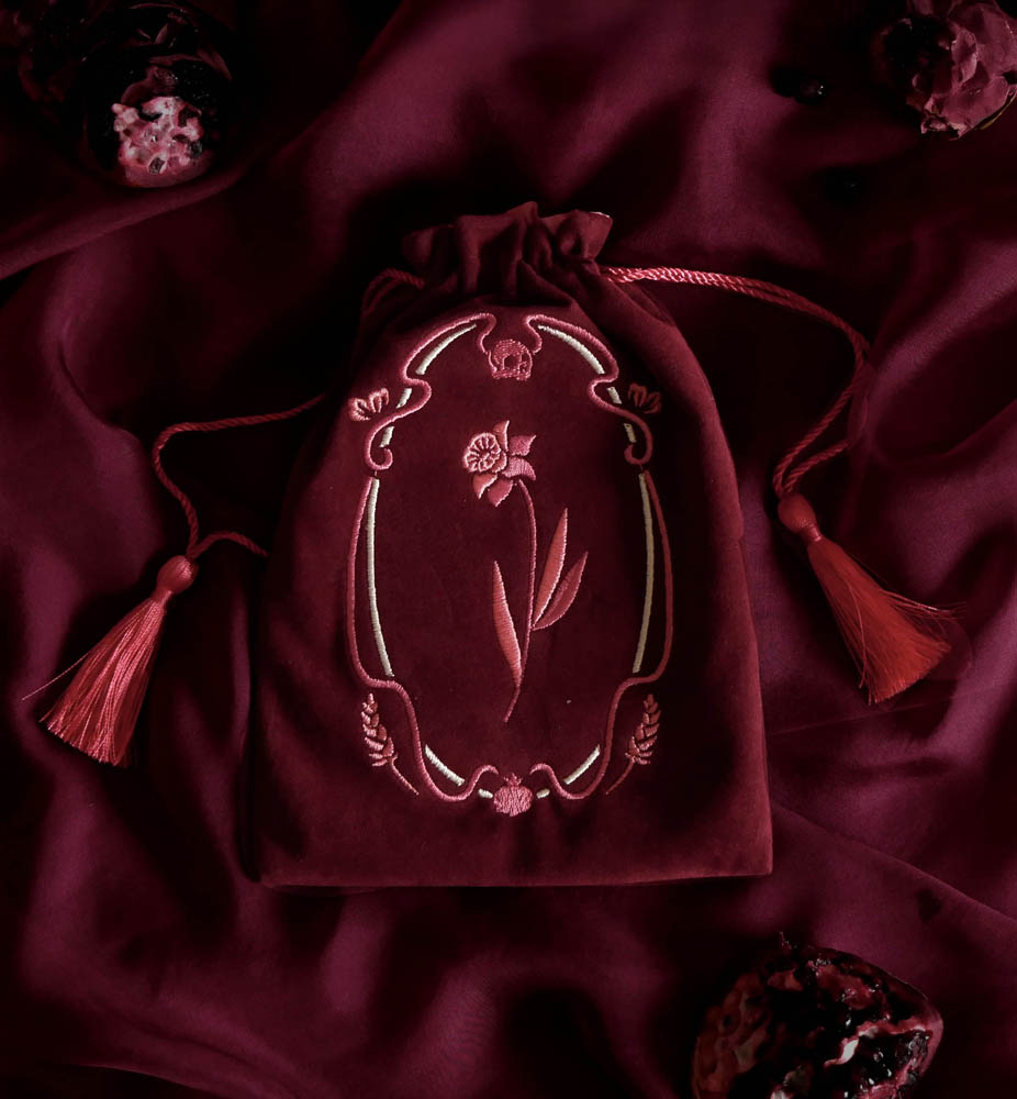 The deep burgundy Persephone velvet drawstring pouch features an embroidered floral design and matching wine coloured tassels. The loot pouch sits on a red fabric background.