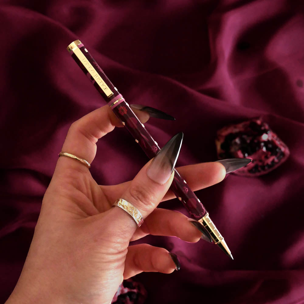 A hand holds the book inspired Persephone pen in wine. it features a dark burgundy body and gold floral accents, the gold pen clip with the words 'Love & Justice' etched in.