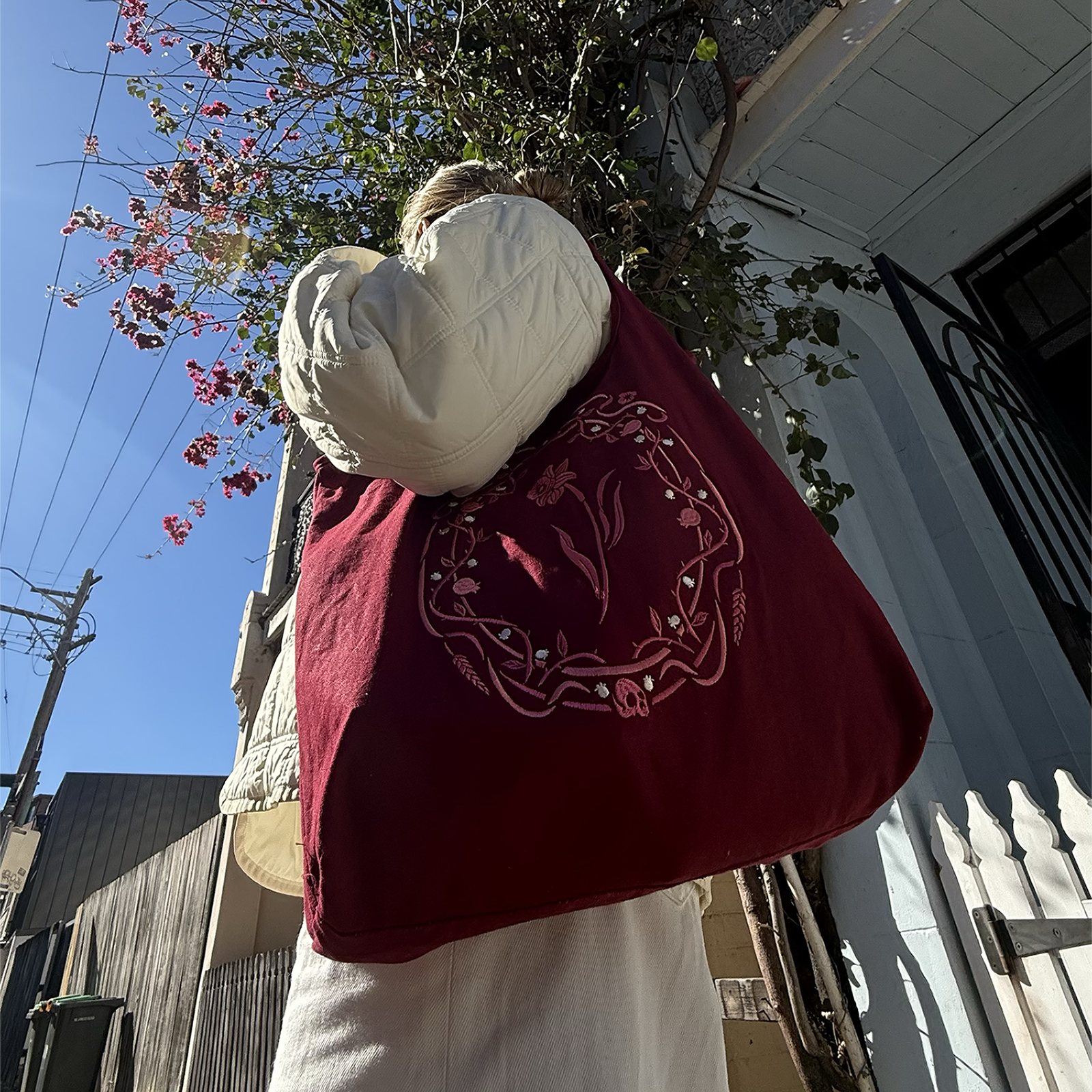 The wine coloured Persephone tote bag features an art nouveau inspired design with flower, skull and pomegranate motifs embroidered in shades of pink and cream. The book inspired tote bag is held by a woman wearing cream in front of a Victorian terrace house.