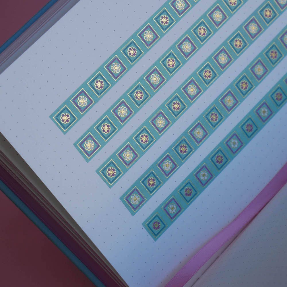 The Poetica washi tape in Antoinette features geometric designs in shades of pink, gold and blue. Three stripes of the tape are in a page of the french inspired notebook against a pink background.