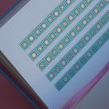 The Poetica washi tape in Antoinette features geometric designs in shades of pink, gold and blue. Three stripes of the tape are in a page of the french inspired notebook against a pink background.