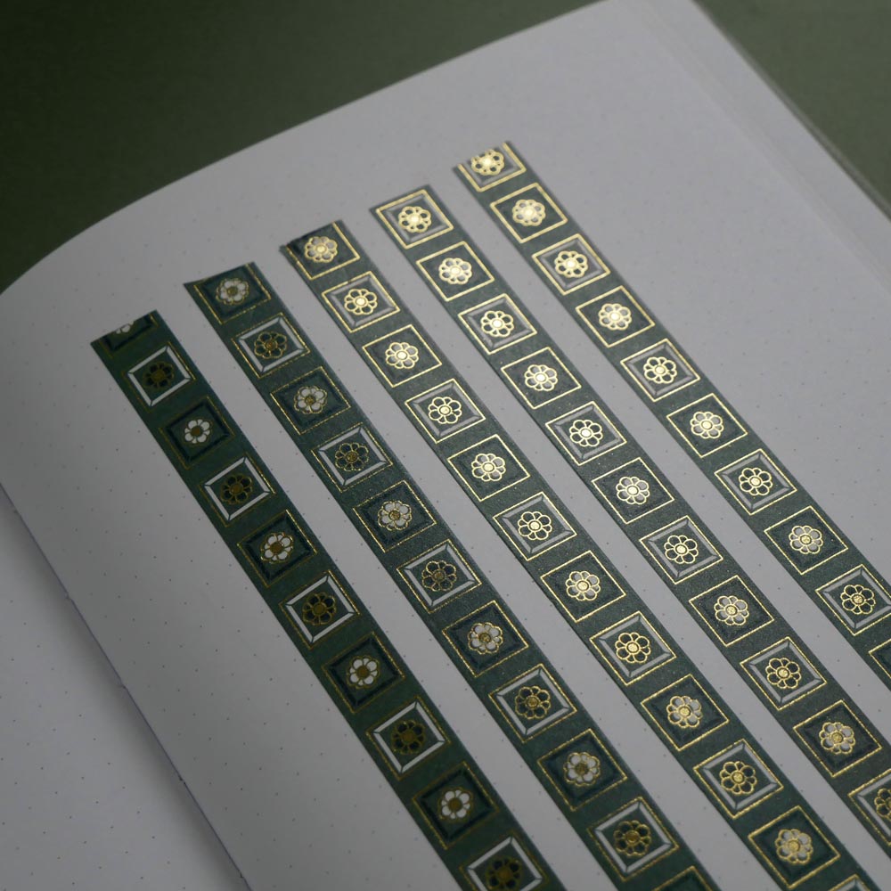 The Sage green variation of the Poetica Literati washi tape. The book inspired washi tape design is shown in five strips on a white journal page sitting on a green background and features geometric flower motifs in shades of dark green, sage green and gold.