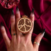 A hand with long ombre nails is holding a The Quirky Cup Collective Clingie (it's like a bookish pop socket or enamel phone grip!) in front of a red fabric background. This romantasy inspired e-reader product is inspired by the story of Persephone and is shaped like a cut pomegranate in shades of red and gold.