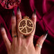 A hand with long ombre nails is holding a The Quirky Cup Collective Clingie (it's like a bookish pop socket or enamel phone grip!) in front of a red fabric background. This romantasy inspired e-reader product is inspired by the story of Persephone and is shaped like a cut pomegranate in shades of red and gold.