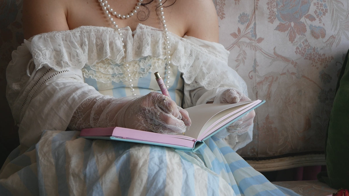 Video of a woman in a blue striped dress with lace gloves writing in a Antionette journal with the matching pen.