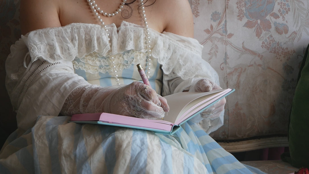 Video of a woman in a blue striped dress with lace gloves writing in a Antionette journal with the matching pen.