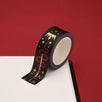 A close up of the Prophecy Foretold washi tape by The Quirky Cup Collective. The roll of fantasy inspired washi tape is on the corner of a white journal on a red background. The bookish washi tape design is black with red and gold detailing- the intricate design showing a crown, red snake, goblet and star icons.