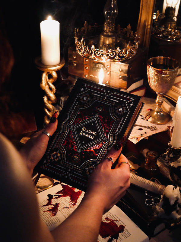 A promo image for the Serpents & Swords collection by The Quirky Cup Collective. A woman is holding a 'Reading Journal' with candles and a crown in the background. The journal is black with gold gilded pages and features red, gold and while foiling with snake and star motifs.