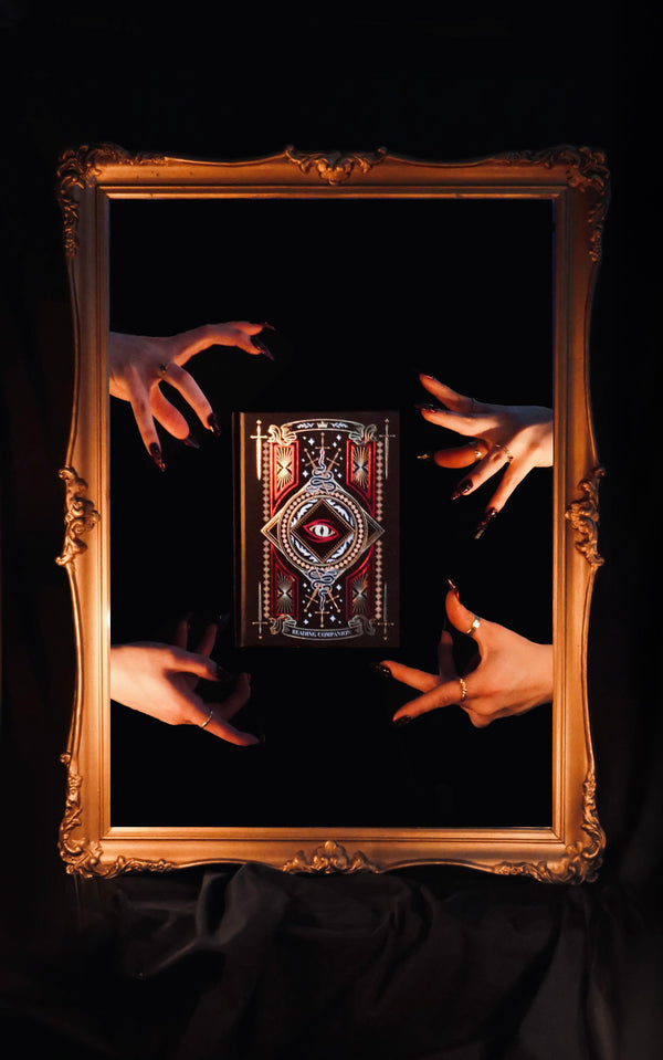 Inside a gold frame, four hands reach out towards a Reading Companion by The Quirky Cup Collective. The intricately foiled journal features serpent and dragon motifs as part of the fantasy book product inspiration.
