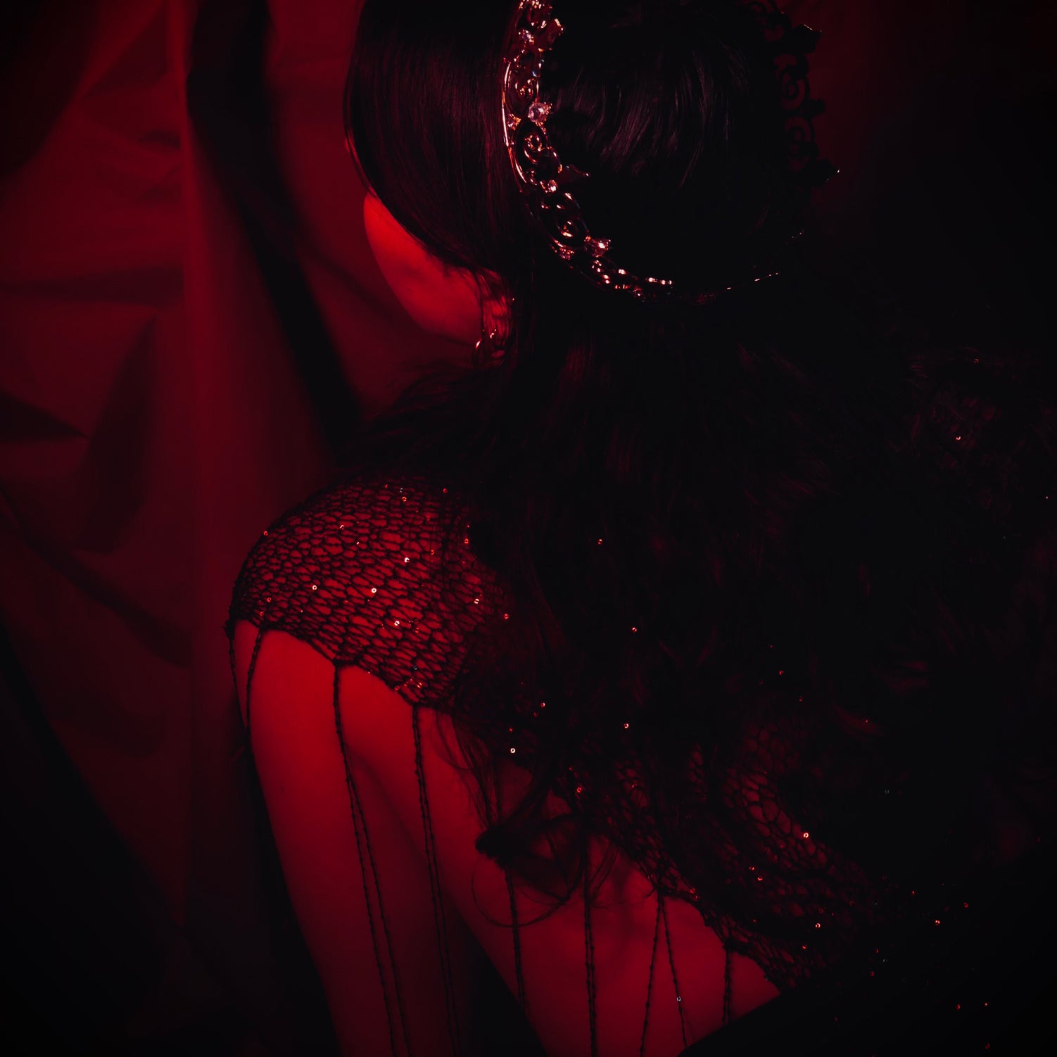 A woman wearing a crown holds and intricate costume is in front of a red curtain, the whole scene tinted red.