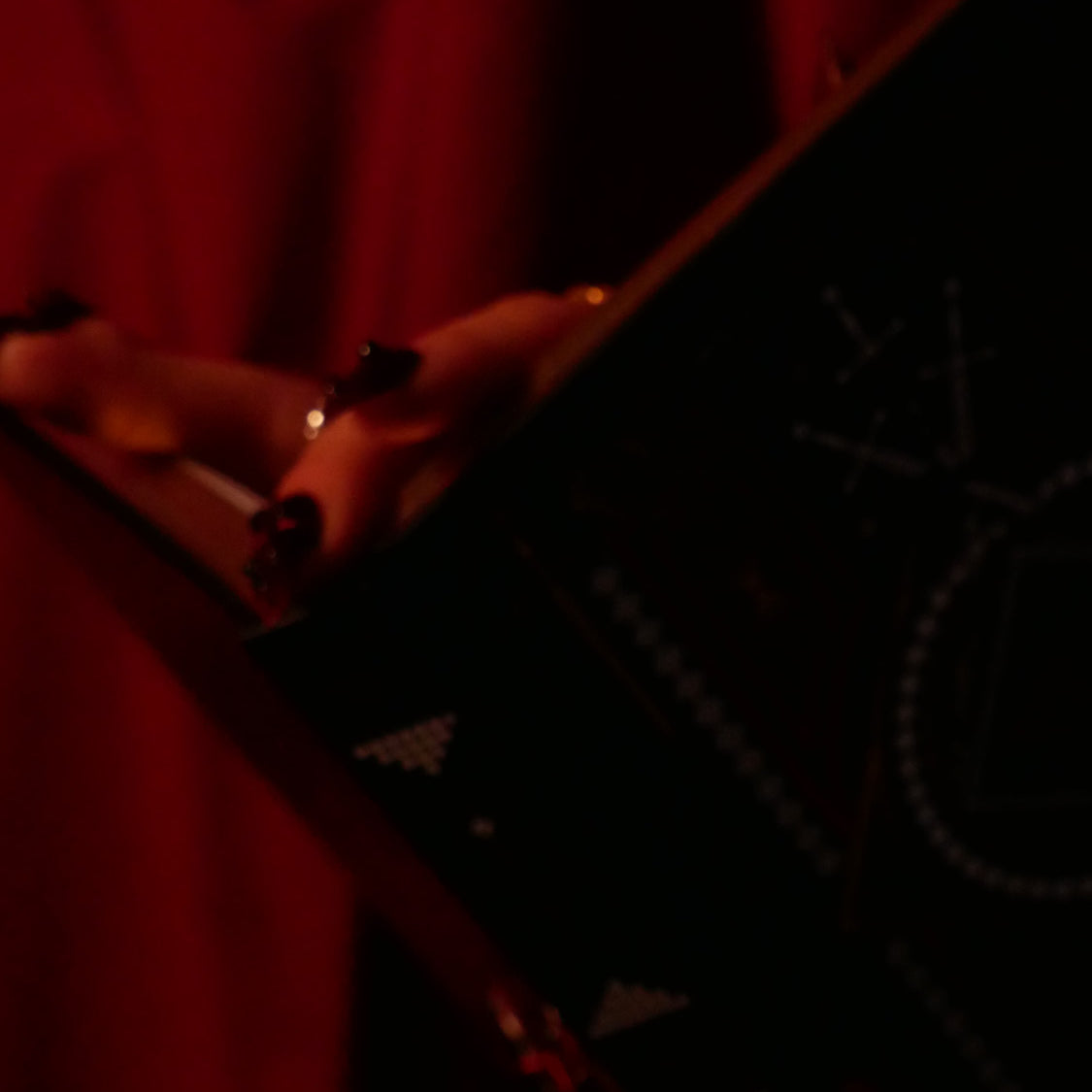 Close-up of a person's hand holding a Serpents & Swords journal in a dark background. The whole image has a deep red colour