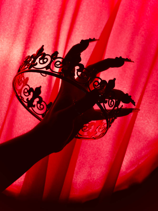 A promo image for Serpents & Swords collection by The Quirky Cup Collective, a Silhouette of a hand with long nails holds a crown in front of a red curtain for a fantasy book aesthetic.
