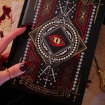 A detail shot of the Quirky Cup Collective 'Destiny Awaits' reading Companion. The black journal features a dragon eye design alongside sword, snake and crown motifs in red, gold and white foils. A hand with intricate red nails holds the journal in front of a stack of handwritten notes covered in blood.