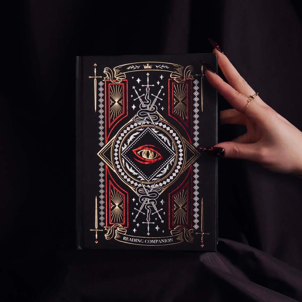 Decorative notebook with fantasy inspired design with swords, crowns and dragon elements held by a hand against a dark background