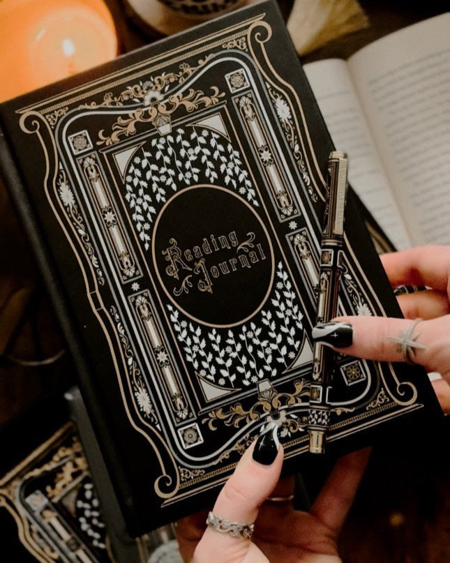 Person holding a black and gold reading journal with decorative cover, surrounded by a cup, candle, and open book.