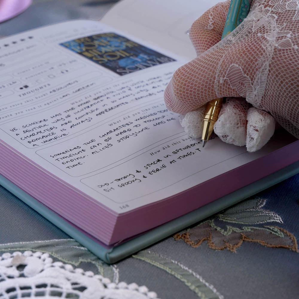 The french inspired reading journal in Antionette colour-way is being written in by a hand wearing lace gloves. The intricate foil journal cover is in shades of pink, gold and blue reference the designs of classic books.