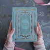 two hands hold the french inspired reading journal in Antionette colour-way over a blue tablecloth. The intricate foil journal cover is in shades of pink, gold and blue reference the designs of classic books making for the perfect victorian inspired journal aesthetic.