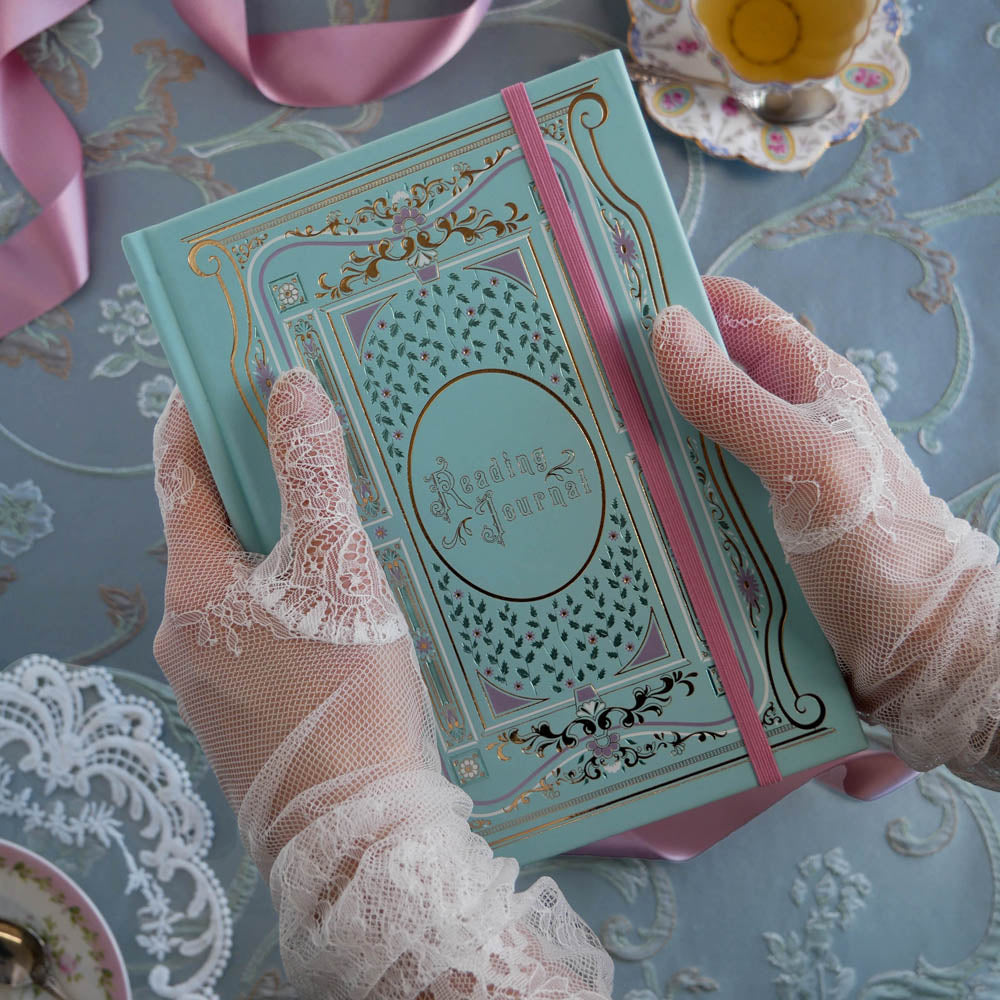 The french inspired reading journal in Antionette colour-way is held by hands wearing lace gloves over a blue tablecloth. The intricate foil notebook cover in shades of pink, gold and blue reference the designs of classic books and the pink elastic closure sits to one side.