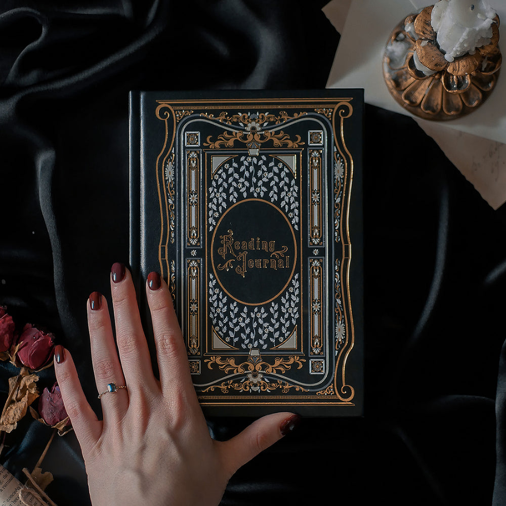 A hand with dark red nail polish and a delicate ring rests on the cover of a Black Reading Journal by The Quirky Cup Collective, showcasing its ornate gold and white embossed design and the words 'Reading Journal' in the center. The journal lies on a smooth black satin fabric, with dried red roses and a decorative candle holder nearby, creating a luxurious and elegant ambiance
