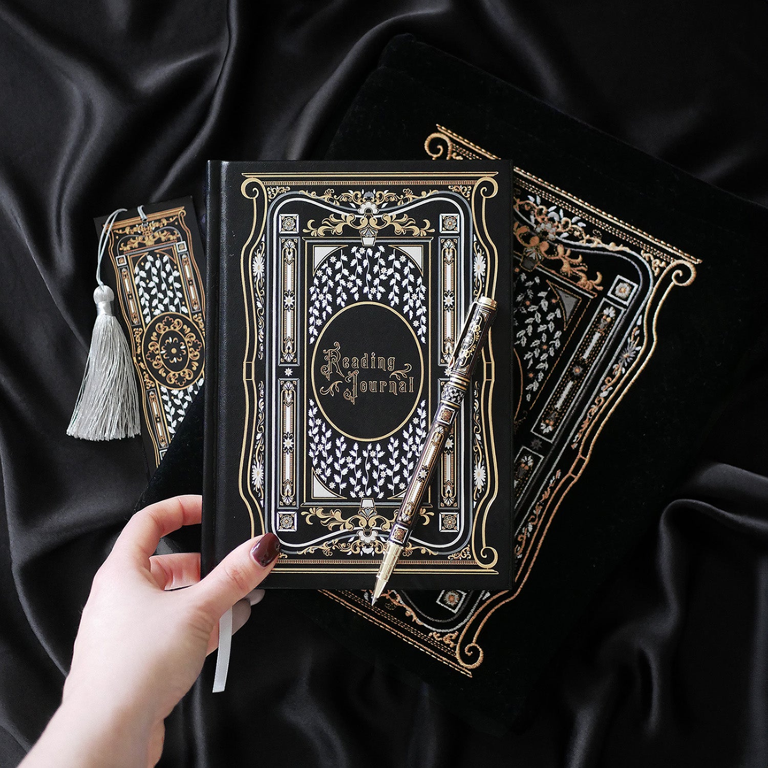 A hand holding a luxurious reading journal with an intricate gold and white design, accompanied by an ornate matching pen. The journal lies on top of a black velvet book sleeve from The Quirky Cup Collective, showcasing a coordinated design. A matching bookmark with a silver tassel is also partially visible. All items are set against a smooth black satin fabric background, creating an elegant and cohesive visual presentation.