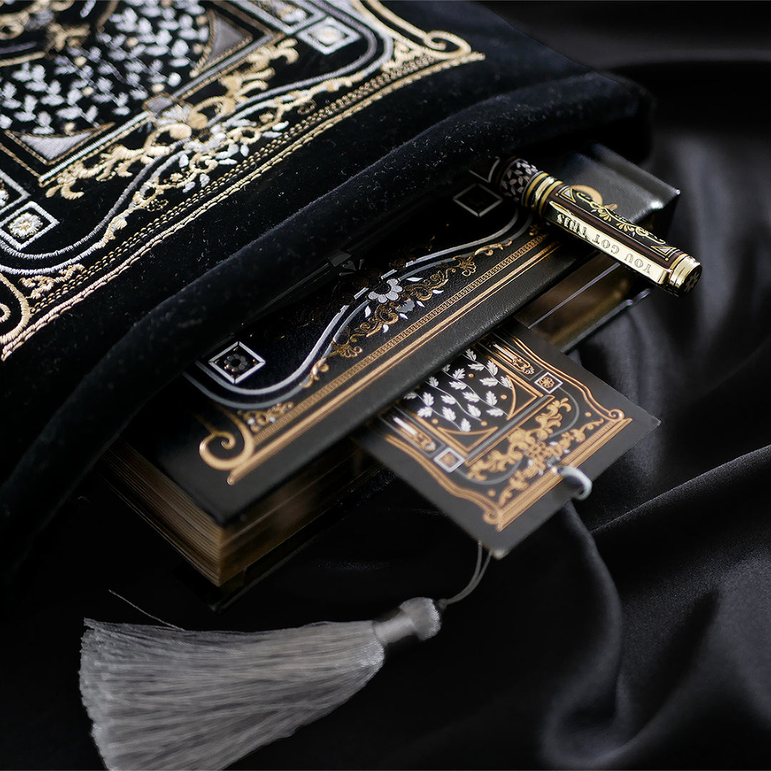 A hand holding a luxurious reading journal with an intricate gold and white design, accompanied by an ornate matching pen. The journal lies on top of a black velvet book sleeve from The Quirky Cup Collective, showcasing a coordinated design. A matching bookmark with a silver tassel is also partially visible. All items are set against a smooth black satin fabric background, creating an elegant and cohesive visual presentation.